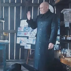 May include: A man in a long, dark gray coat waves in a rustic, wooden interior. The coat has a button-down front and a wide collar. Behind him are drawings and shelves with jars and containers.