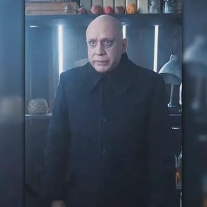 May include: A man with a shaved head wearing a black coat with a large collar. The coat is buttoned up, and the man is looking directly at the camera. The background includes a shelf with various items.