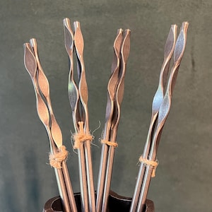 Puede incluir: Primer plano de un juego de cuatro bombillas de metal para yerba mate, cada una con un tallo retorcido y un filtro en forma de hoja. Las bombillas están atadas con hilo y están en un recipiente de metal oscuro, sobre un fondo verde.