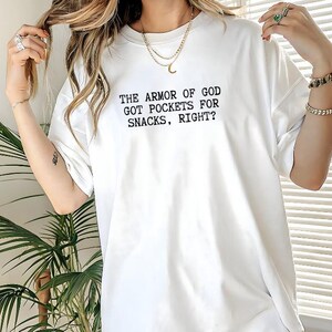 Puede incluir: Camiseta blanca con el texto negro: "THE ARMOR OF GOD GOT POCKETS FOR SNACKS, RIGHT?" La camiseta tiene mangas cortas, cuello redondo y un corte holgado.
