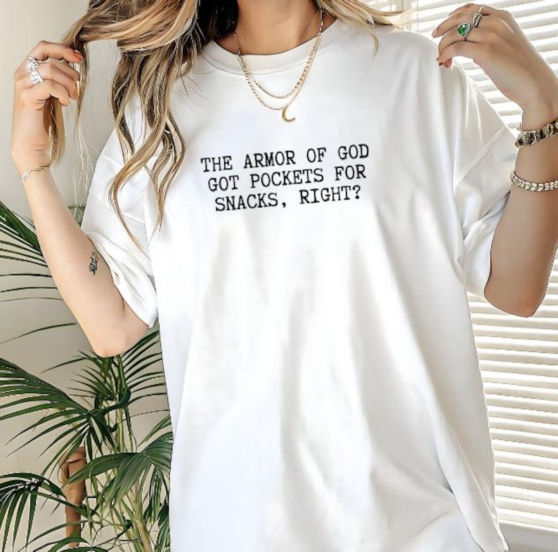 Puede incluir: Camiseta blanca con el texto negro: "THE ARMOR OF GOD GOT POCKETS FOR SNACKS, RIGHT?" La camiseta tiene mangas cortas, cuello redondo y un corte holgado.