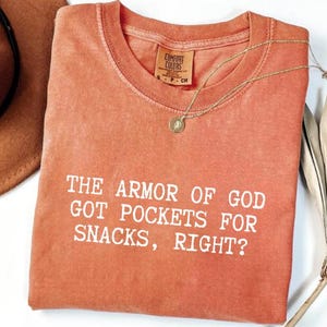 Puede incluir: Una camiseta Comfort Colors de color coral con la frase "THE ARMOR OF GOD GOT POCKETS FOR SNACKS, RIGHT?" en letras blancas. Un collar dorado con un colgante circular descansa sobre la camiseta. Un sombrero marr&oacute;n y plumas blancas en el fondo.