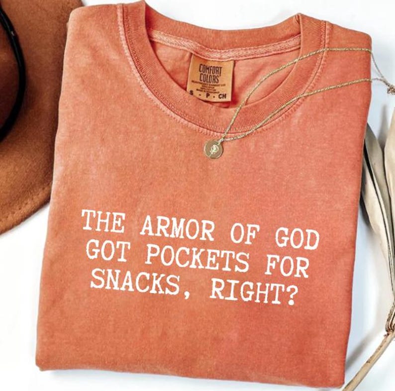 Puede incluir: Una camiseta Comfort Colors de color coral con la frase "THE ARMOR OF GOD GOT POCKETS FOR SNACKS, RIGHT?" en letras blancas. Un collar dorado con un colgante circular descansa sobre la camiseta. Un sombrero marr&oacute;n y plumas blancas en el fondo.