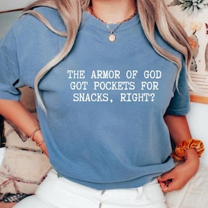Puede incluir: Camiseta azul claro con el texto blanco: "THE ARMOR OF GOD GOT POCKETS FOR SNACKS, RIGHT?". La camiseta tiene mangas cortas y cuello redondo.