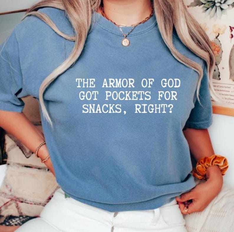 Puede incluir: Camiseta azul claro con el texto blanco: "THE ARMOR OF GOD GOT POCKETS FOR SNACKS, RIGHT?". La camiseta tiene mangas cortas y cuello redondo.