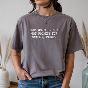 Puede incluir: Una camiseta gris con el texto blanco: "THE ARMOR OF GOD GOT POCKETS FOR SNACKS, RIGHT?" La camiseta es de corte holgado y se lleva con unos vaqueros azules. La imagen es un primer plano de la camiseta y de la persona que la lleva.