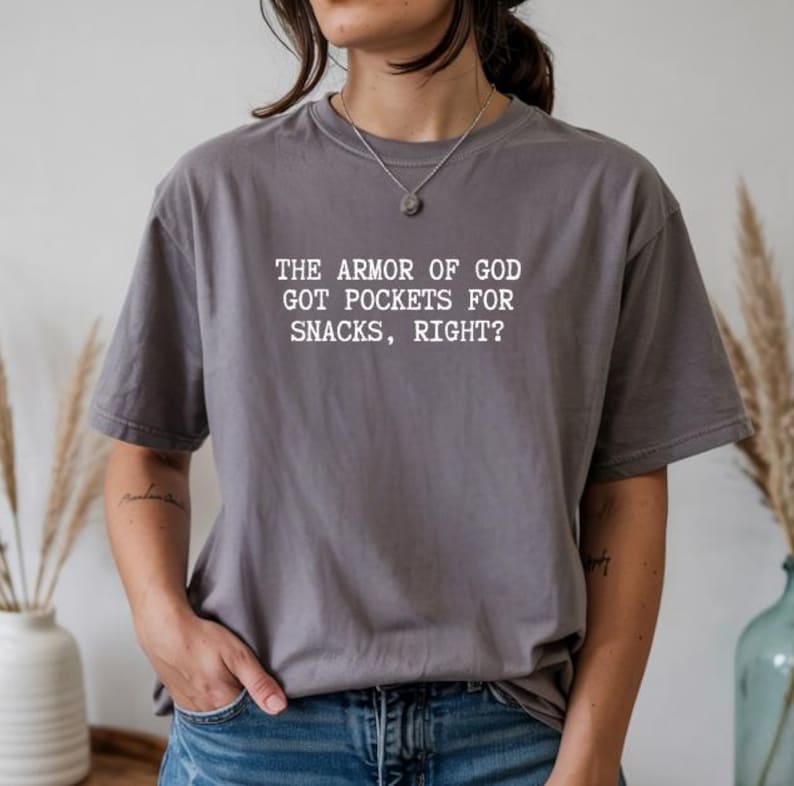 Puede incluir: Una camiseta gris con el texto blanco: "THE ARMOR OF GOD GOT POCKETS FOR SNACKS, RIGHT?" La camiseta es de corte holgado y se lleva con unos vaqueros azules. La imagen es un primer plano de la camiseta y de la persona que la lleva.