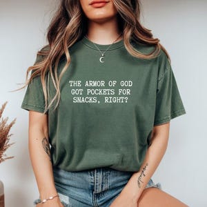 Puede incluir: Una camiseta verde de manga corta con el texto blanco "THE ARMOR OF GOD GOT POCKETS FOR SNACKS, RIGHT?". La camiseta tiene un corte relajado y est&aacute; hecha de una tela suave. La camiseta es de color liso.