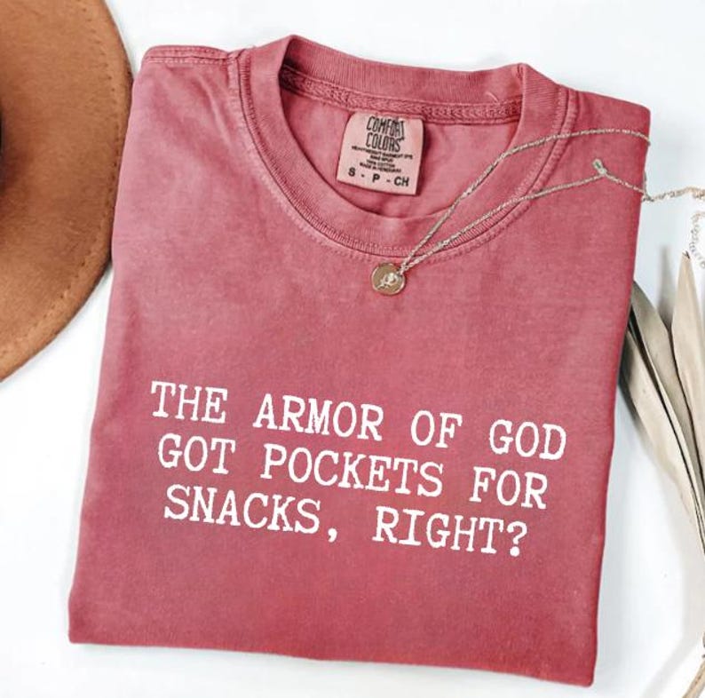 Puede incluir: Una camiseta Comfort Colors rosa descolorido con el texto blanco "THE ARMOR OF GOD GOT POCKETS FOR SNACKS, RIGHT?". Un collar plateado con un colgante est&aacute; drapeado sobre la camisa. La camisa est&aacute; doblada sobre una superficie blanca.