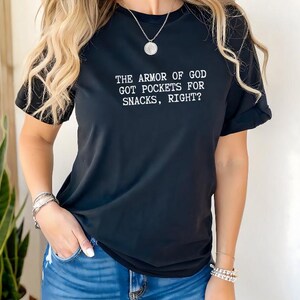 Puede incluir: Camiseta negra de manga corta con el texto blanco "THE ARMOR OF GOD GOT POCKETS FOR SNACKS, RIGHT?". La camiseta se combina con unos vaqueros azules. La modelo lleva pulseras plateadas y un collar.