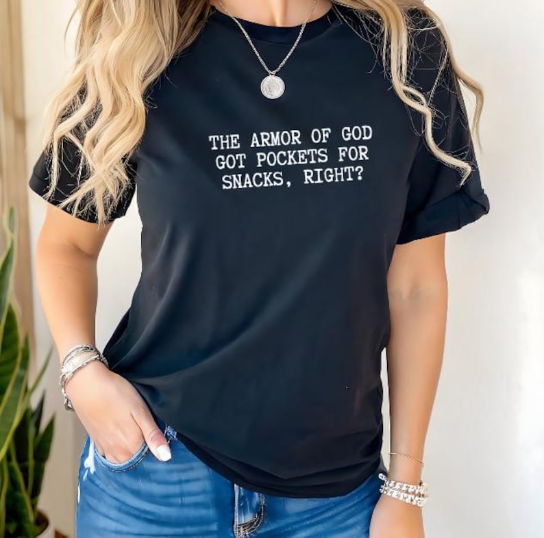 Puede incluir: Camiseta negra de manga corta con el texto blanco "THE ARMOR OF GOD GOT POCKETS FOR SNACKS, RIGHT?". La camiseta se combina con unos vaqueros azules. La modelo lleva pulseras plateadas y un collar.
