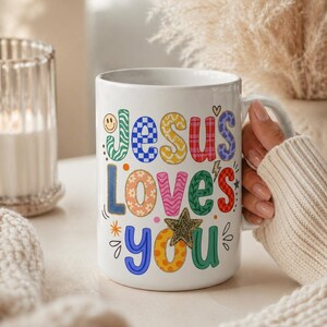 May include: White ceramic mug with the text "Jesus Loves You" in colorful, playful lettering. The mug features a smiley face, stars, and a heart design. The mug is held by a person wearing a cream-colored sweater.