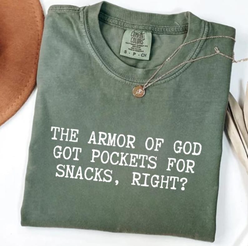 Puede incluir: Camiseta verde oliva Comfort Colors con el texto blanco "THE ARMOR OF GOD GOT POCKETS FOR SNACKS, RIGHT?". Un collar dorado con un colgante descansa sobre la camiseta.