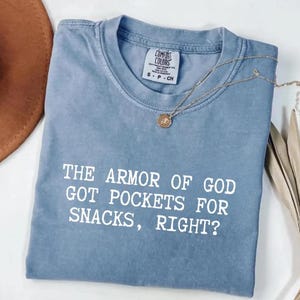 Puede incluir: Una camiseta azul Comfort Colors con el texto "THE ARMOR OF GOD GOT POCKETS FOR SNACKS, RIGHT?" en blanco. Un collar dorado con un colgante est&aacute; sobre la camiseta. Un sombrero marr&oacute;n es visible en la esquina superior izquierda.
