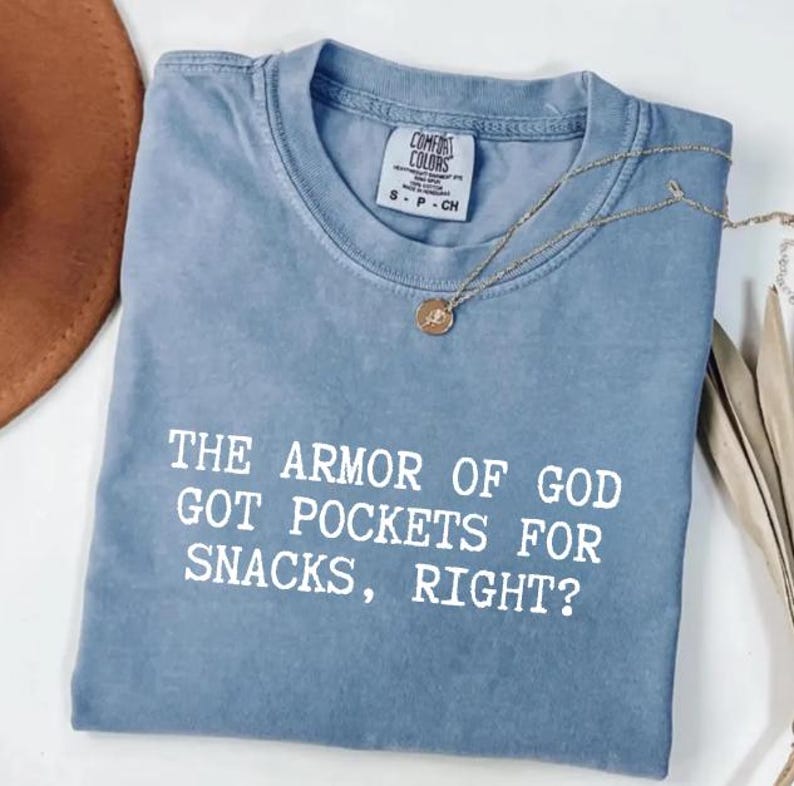 Puede incluir: Una camiseta azul Comfort Colors con el texto "THE ARMOR OF GOD GOT POCKETS FOR SNACKS, RIGHT?" en blanco. Un collar dorado con un colgante est&aacute; sobre la camiseta. Un sombrero marr&oacute;n es visible en la esquina superior izquierda.