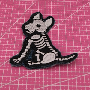 Op de afbeelding: Geborduurde patch met een zwart honden skelet ontwerp met witte details. De patch heeft een donkerblauwe rand en is geplaatst tegen een roze raster achtergrond. Ideaal voor jassen, tassen of andere stoffen items.