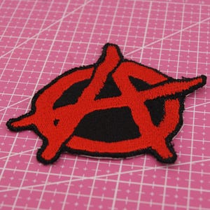 Puede incluir: Parche bordado con el símbolo de la anarquía, una "A" roja dentro de un círculo negro. El parche tiene un contorno rojo y está sobre un fondo rosa cuadriculado. Ideal para personalizar ropa o accesorios.