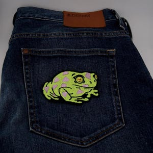 Könnte beinhalten: Dunkelblaue Jeans mit einem Flicken einer Frosch-Applikation auf der Gesäßtasche. Der Flicken ist limettengrün mit rosa Punkten und schwarzer Umrandung. Ein braunes Lederetikett mit dem Text "& DENIM" ist sichtbar.