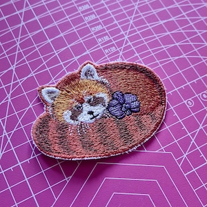 May include: Embroidered patch featuring a red panda curled up with a purple flower. The patch has detailed stitching in shades of brown, orange, white, and purple. The patch is on a pink cutting mat.