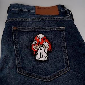 Peut inclure: Un jean en denim bleu foncé avec un écusson champignon rouge et blanc sur la poche arrière. L'écusson présente un design de style dessin animé avec un chapeau rouge, des taches blanches et une tige blanche. Le jean a un patch en cuir marron avec le texte "& DENIM".