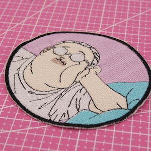 May include: Embroidered patch depicting a cartoon character with glasses, resting their chin on their hand. The patch has a pink and blue colour scheme, with a black border. The background is a pink cutting mat.