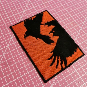 Może przedstawiać: Prostokątna naszywka z pomarańczowym tłem i czarnym wzorem kruka. Kruk jest przedstawiony w sylwetce, ze skrzydłami rozłożonymi. Naszywka ma czarną obwódkę i jest prawdopodobnie wykonana z tkaniny.