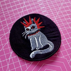 May include: A round, black embroidered patch featuring a gray cat with a red mohawk and collar. The cat is sitting with its tail curled. The patch is on a pink cutting mat.