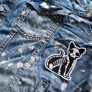 May include: A blue denim jacket with white paint splatters and a black and white cat skeleton patch. The jacket has a classic design with button closures and pockets, perfect for casual wear.