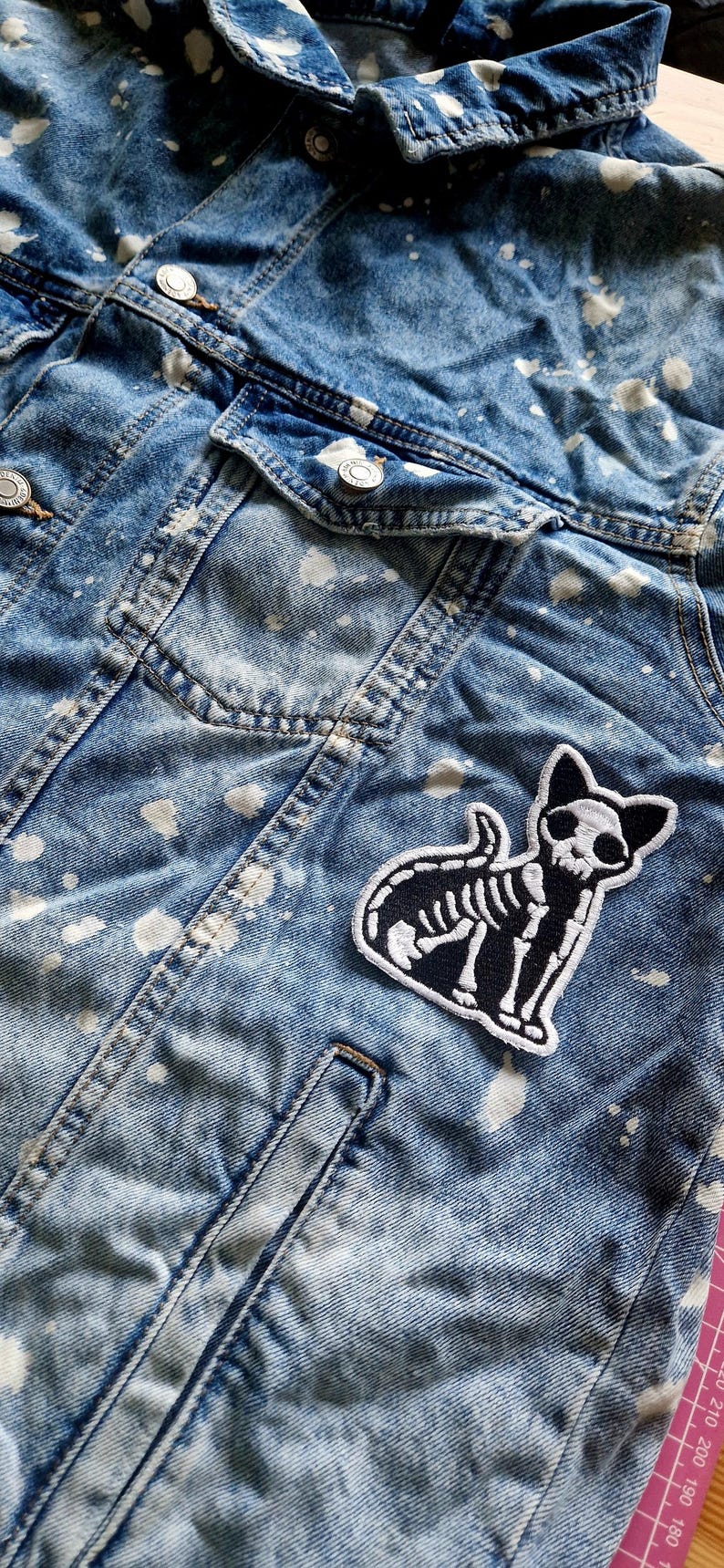 May include: A blue denim jacket with white paint splatters and a black and white cat skeleton patch. The jacket has a classic design with button closures and pockets, perfect for casual wear.