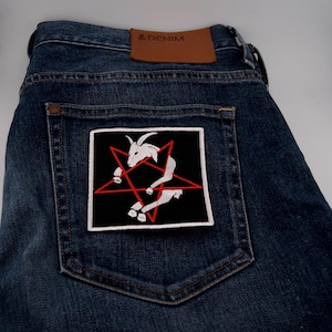 Può includere: Jeans blu scuro con una toppa quadrata raffigurante una capra bianca all'interno di un pentagramma rosso su sfondo nero. La toppa è sulla tasca posteriore. I jeans hanno un'etichetta in pelle marrone.