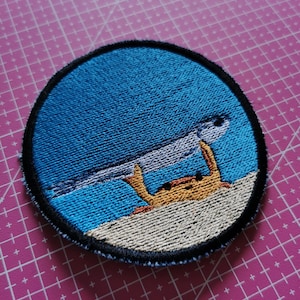 May include: Embroidered patch featuring a crab holding a fish in a blue ocean scene. The crab is orange and the fish is silver. The patch has a black border and is circular in shape.