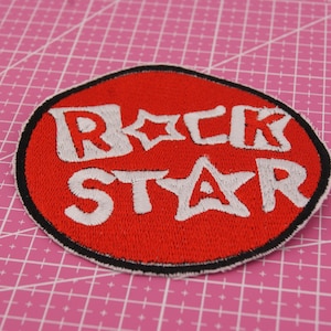 Può includere: Una toppa rossa di forma ovale con le parole "ROCK STAR" ricamate in bianco, bordate di nero. La toppa ha un bordo nero ed è posta su uno sfondo a griglia rosa. Le lettere includono dettagli a forma di stella.