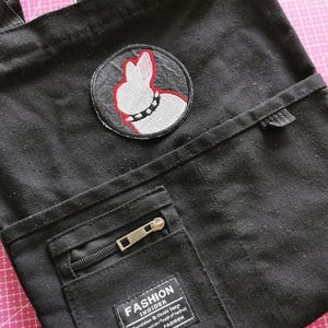 May include: Black canvas tote bag with a circular patch featuring a white rabbit design with a red outline and a studded collar. The bag has a front pocket with a zipper and a label that reads "FASHION INSIDER".