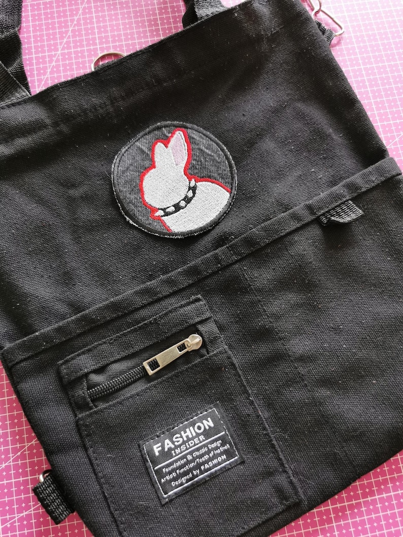 May include: Black canvas tote bag with a circular patch featuring a white rabbit design with a red outline and a studded collar. The bag has a front pocket with a zipper and a label that reads "FASHION INSIDER".