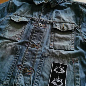 May include: A light blue denim jacket with multiple pockets and button closures. The jacket features a black patch with a white eye design. Red stitching accents the seams and pockets. The jacket is made of a durable denim material.