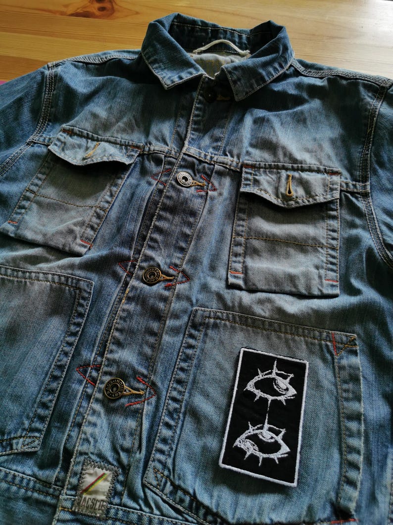 May include: A light blue denim jacket with multiple pockets and button closures. The jacket features a black patch with a white eye design. Red stitching accents the seams and pockets. The jacket is made of a durable denim material.