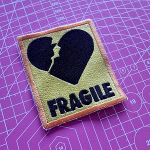 May include: Embroidered patch with a black broken heart design on a yellow background. The word "FRAGILE" is printed in bold black letters below the heart. The patch has an orange border and is set against a purple cutting mat.