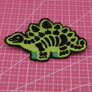 May include: Embroidered patch with a green and black dinosaur design. The dinosaur has a detailed outline with black spots and a black spine. The patch is on a pink cutting mat.