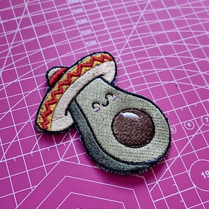 Può includere: Toppa ricamata con un avocado sorridente che indossa un sombrero. L'avocado è verde con un nocciolo marrone e dettagli ricamati. Il sombrero è beige con accenti rossi e gialli. Una toppa divertente a tema cibo.