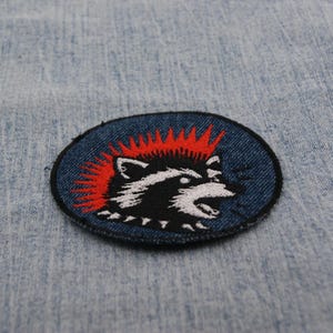 May include: Oval patch featuring a raccoon with a red mohawk and a black and white face. The raccoon is set against a dark blue background with a black border. The patch is likely for clothing or accessories.