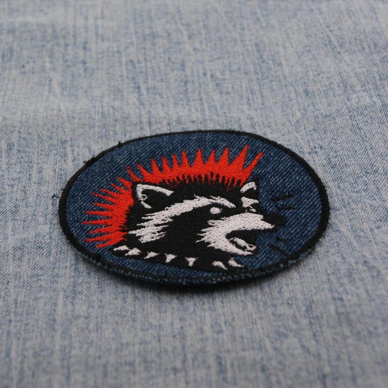 May include: Oval patch featuring a raccoon with a red mohawk and a black and white face. The raccoon is set against a dark blue background with a black border. The patch is likely for clothing or accessories.