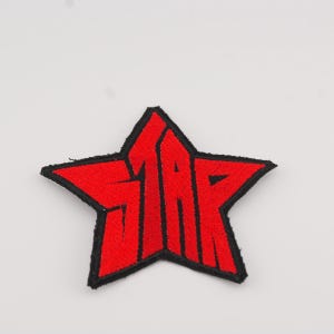 Puede incluir: Parche bordado en forma de estrella roja con la palabra "STAR" en letras blancas estilizadas. El parche tiene un borde negro y está diseñado para su aplicación en ropa o accesorios.
