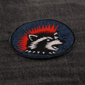 May include: An embroidered patch featuring a raccoon with a red mohawk and open mouth, set against a blue oval background. The raccoon is black and white, with a detailed design. The patch is on a dark gray denim fabric.