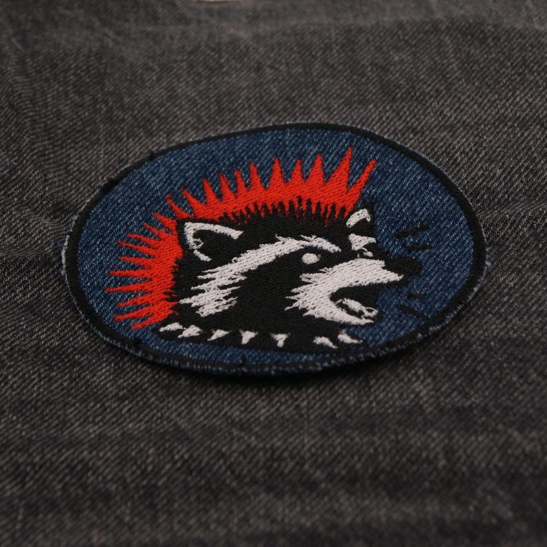 May include: An embroidered patch featuring a raccoon with a red mohawk and open mouth, set against a blue oval background. The raccoon is black and white, with a detailed design. The patch is on a dark gray denim fabric.