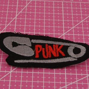 Könnte beinhalten: Gestickter Aufnäher in Form einer Sicherheitsnadel mit dem Wort "PUNK" in roten Buchstaben. Der Aufnäher hat einen schwarzen Rand und graue und weiße Nähte. Der Hintergrund ist eine rosa Schneidematte.