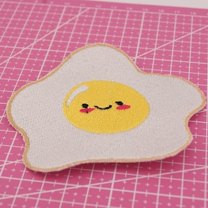 Op de afbeelding: Geborduurde patch in de vorm van een gebakken ei met een lachend gezicht. Het eiwit is gebroken wit, de dooier is geel en het gezicht heeft een zwarte mond en roze wangen. De patch ligt op een roze snijmat.