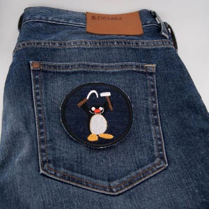 May include: Blue denim jeans with a cartoon penguin patch on the back pocket. The patch features a white penguin with a red nose and orange feet, holding tools. A brown leather patch with "& DENIM" is visible.