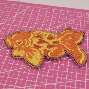 May include: Embroidered goldfish patch in orange and yellow, with a denim blue border. The fish design features detailed scales and fins. Ideal for adding to clothing or accessories. The patch is approximately 10 cm in length.