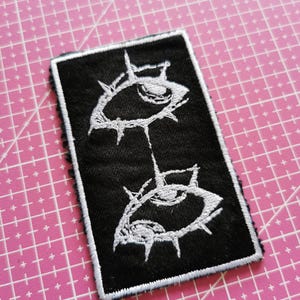 Könnte beinhalten: Schwarzer rechteckiger Aufnäher mit einem weißen, gestickten Design von zwei stilisierten Augen, die durch eine Linie verbunden sind. Die Augen haben einen stacheligen Rand. Der Aufnäher hat einen weißen Rand.
