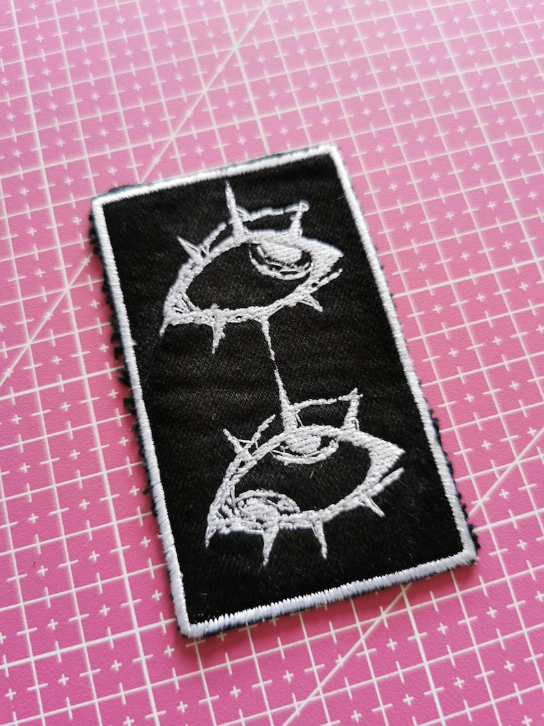 May include: Black rectangular patch with a white embroidered design of two stylized eyes connected by a line. The eyes have a spiky, thorn-like border. The patch has a white border and is on a pink grid surface.